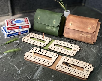 May include: A wooden cribbage board with a leather carrying case. The case is brown with the word "Sebas" and a smiley face on it. The other case is green with the words "crit shit" on it. The cribbage board has a slot for cards and pegs.