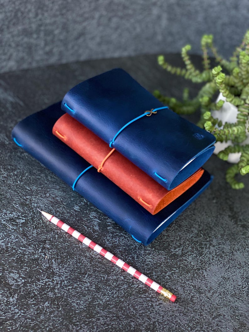 May include: A stack of three leather journals in navy blue and rust colors, secured with elastic cords. A red and white striped pencil rests in the foreground. The journals are stacked on a dark, textured surface.