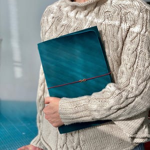 May include: Teal-colored leather portfolio with a red elastic closure. The portfolio is held against a person wearing a beige cable-knit sweater. The portfolio has a diagonal striped pattern.