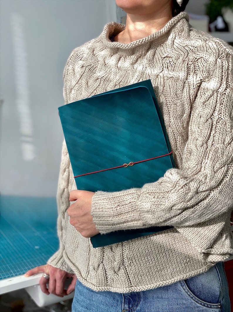 May include: Teal-colored leather portfolio with a red elastic closure. The portfolio is held against a person wearing a beige cable-knit sweater. The portfolio has a diagonal striped pattern.