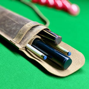 May include: A brown leather case with a strap for carrying pool cues. The case is open and three cues are inside. The cues are a white cue, a black cue, and a brown cue.