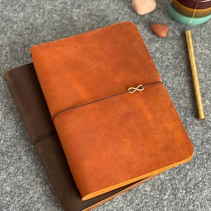 Puede incluir: Se muestran dos diarios de cuero, uno en marrón oscuro y otro en un tono naranja cálido. El diario naranja presenta un cierre con un símbolo de infinito dorado. Los diarios están cerrados y parecen nuevos.