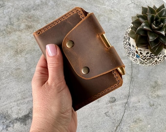 May include: A brown leather wallet with a brass clasp and two brass rivets. The wallet is held in a hand. The stitching is visible along the edges. A small succulent plant in a mosaic pot is in the background.
