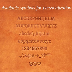 May include: A brown leather surface with a list of available symbols for personalization. The symbols include uppercase and lowercase letters, numbers, punctuation marks, and a few emojis.