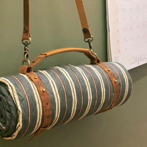 May include: A striped, quilted blanket with a brown leather strap and handle. The blanket is rolled up and hanging from a hook on a wall.