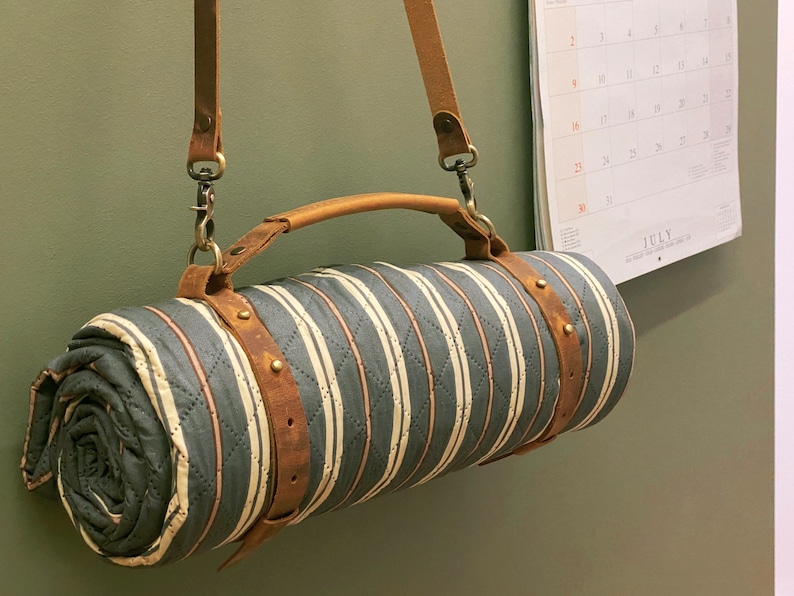 May include: A striped, quilted blanket with a brown leather strap and handle. The blanket is rolled up and hanging from a hook on a wall.