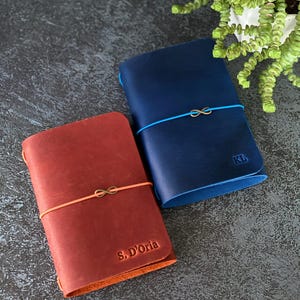 May include: Two leather journals, one red and one blue, are displayed. The red journal has the text "S. D'Oria" embossed on the cover, and the blue journal has the initials "KL". Both journals are secured with elastic bands and feature an infinity symbol charm.