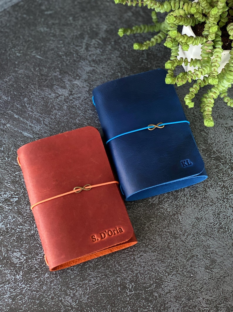May include: Two leather journals, one red and one blue, are displayed. The red journal has the text "S. D'Oria" embossed on the cover, and the blue journal has the initials "KL". Both journals are secured with elastic bands and feature an infinity symbol charm.