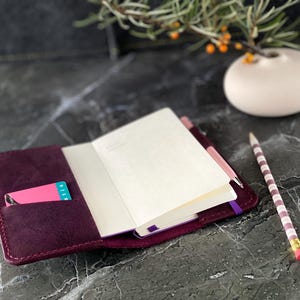 May include: A burgundy leather notebook cover with a pink card holder and a purple elastic closure. The notebook is open and has blank white pages. A striped pencil is lying on the table next to the notebook.