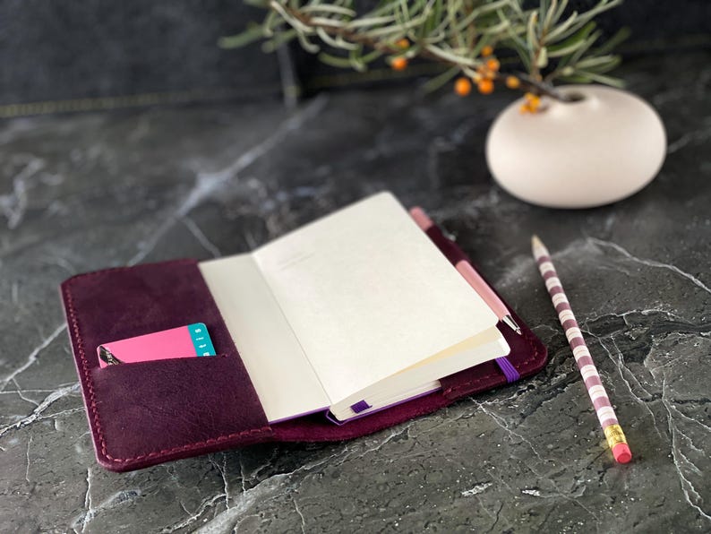 May include: A burgundy leather notebook cover with a pink card holder and a purple elastic closure. The notebook is open and has blank white pages. A striped pencil is lying on the table next to the notebook.