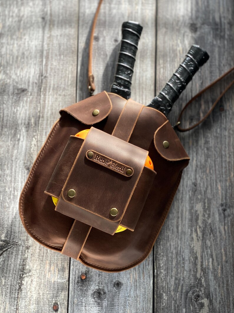 Puede incluir: Una bolsa de cuero marr&oacute;n con una etiqueta de nombre personalizada que dice "Rene' Bird". La bolsa tiene dos raquetas de paddleball negras en su interior y una pelota de paddleball amarilla.