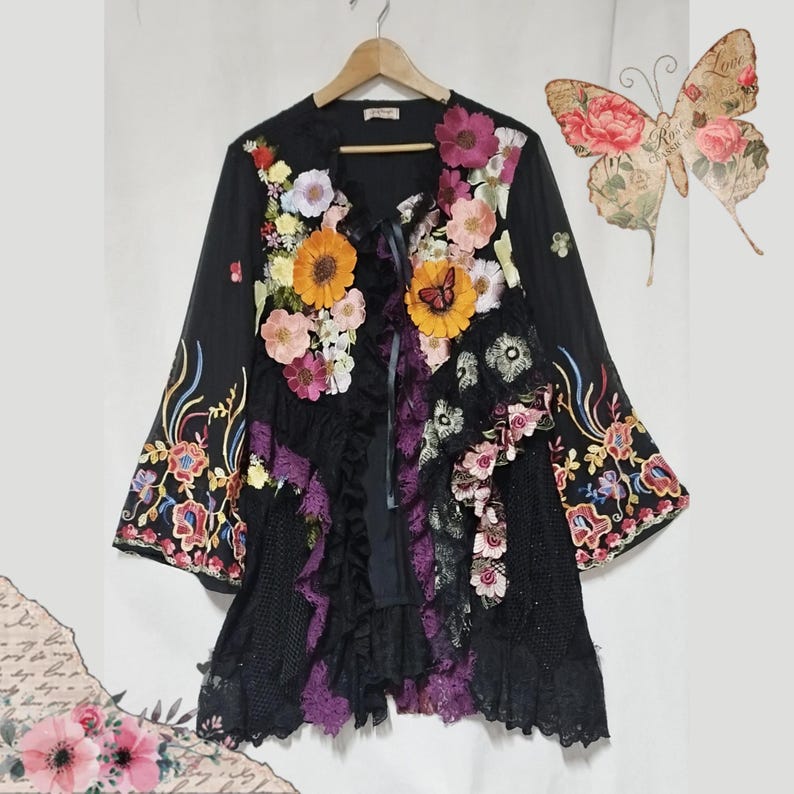 May include: A black long-sleeve jacket featuring a front panel adorned with various colorful fabric flowers, including orange, pink, and white blossoms. The garment includes black lace ruffles, embroidered floral sleeve cuffs, and a butterfly graphic.