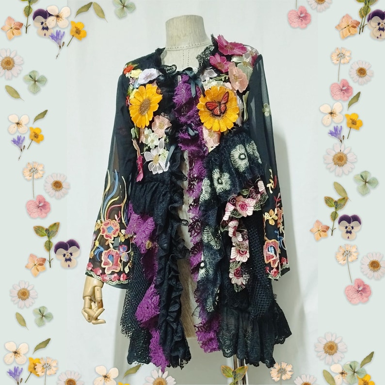 May include: A black long-sleeve jacket featuring layered ruffles, purple lace trim, and various floral appliques including yellow sunflowers and a butterfly. The garment has a sheer fabric base with intricate embroidery and decorative ribbons.
