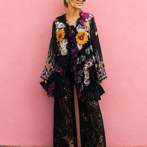 May include: A black long-sleeved jacket featuring vibrant floral embroidery, ruffles, and a butterfly motif, paired with wide-leg black lace trousers. The jacket has a dark sheer base with colorful flower appliqu&eacute;s and black lace trim details.