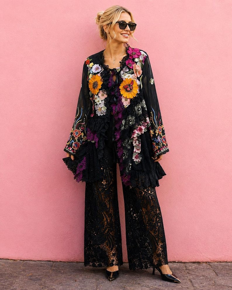 May include: A black long-sleeved jacket featuring vibrant floral embroidery, ruffles, and a butterfly motif, paired with wide-leg black lace trousers. The jacket has a dark sheer base with colorful flower appliqu&eacute;s and black lace trim details.