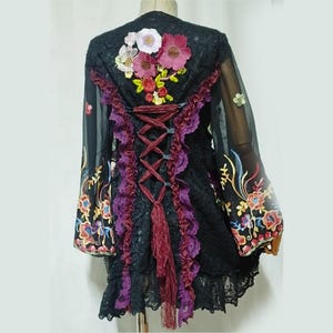 May include: A black lace jacket featuring long sheer sleeves with colorful floral embroidery. The back displays a central corset-style lace-up closure with deep purple ruffled trim, accented by a cluster of embroidered flowers at the top.