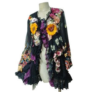 May include: A black long-sleeve jacket featuring layered ruffles, lace trim, and vibrant floral appliques in shades of yellow, pink, and orange. The garment includes a butterfly accent on a sunflower patch and intricate embroidery on the sleeves.