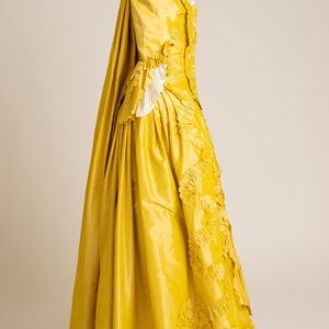May include: A yellow silk gown with a long train and a fitted bodice. The gown is decorated with large, ruffled flowers.