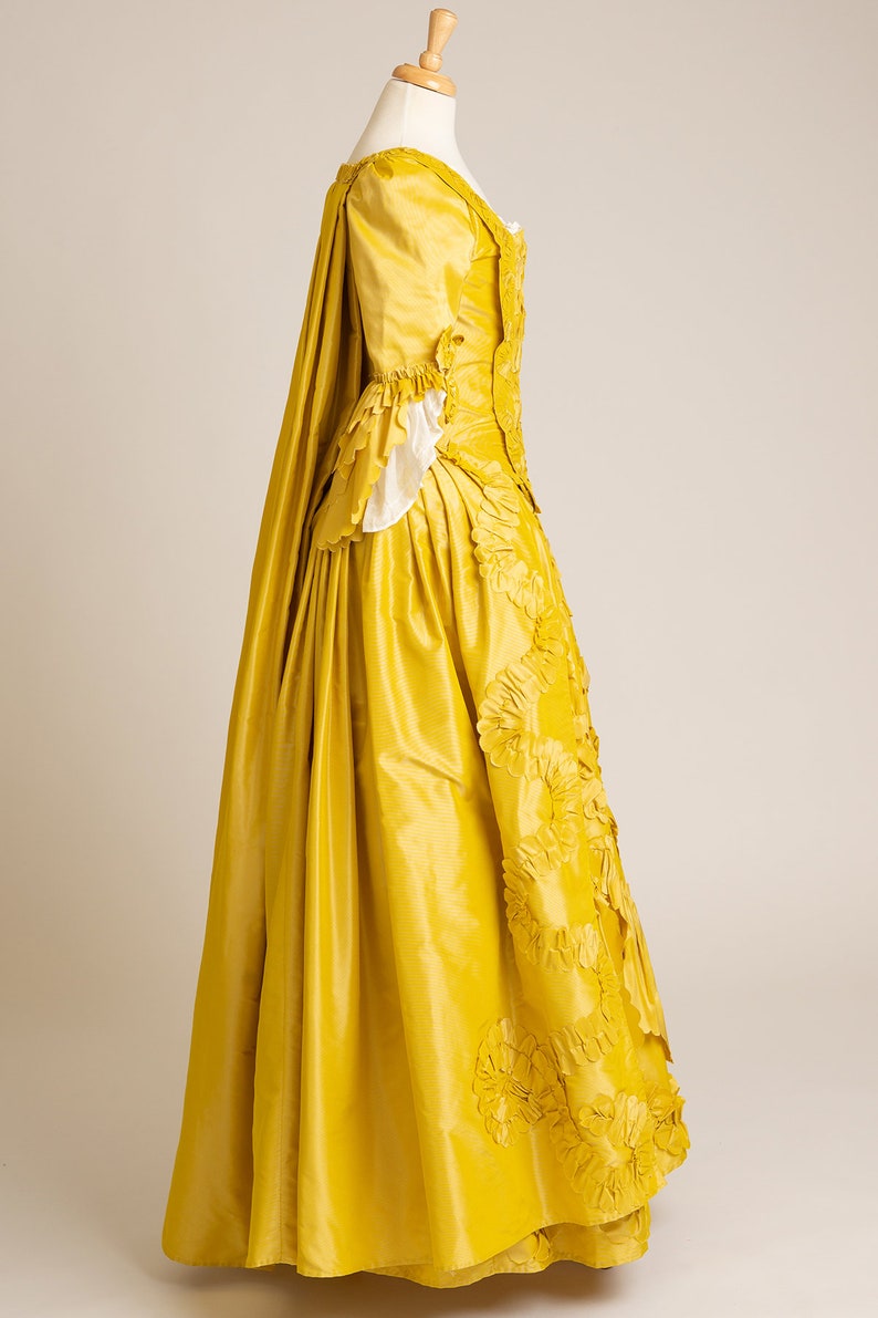 May include: A yellow silk gown with a long train and a fitted bodice. The gown is decorated with large, ruffled flowers.