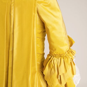 May include: A yellow silk dress with a ruffled sleeve. The dress has a square neckline and a fitted bodice. The skirt is full and flowing.