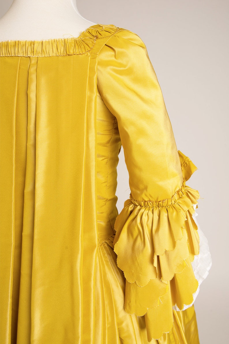 May include: A yellow silk dress with a ruffled sleeve. The dress has a square neckline and a fitted bodice. The skirt is full and flowing.