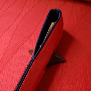 May include: A red fabric case with a navy blue interior and trim. The case is partially open, revealing a glimpse of a gold-colored object inside. The case is resting on a dark brown object, and the background is a textured red surface.