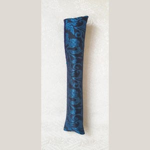 May include: A long, narrow, cylindrical fabric tube featuring a deep black base color with a vibrant, shimmering teal blue floral damask pattern woven throughout the surface. The item has a straight, elongated shape with a flat, angled top edge.