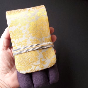 May include: A rectangular, gold and cream-coloured fabric case with a floral pattern, secured by a striped band. The case is held over a purple fabric. The case is likely for glasses or a phone.