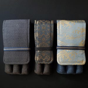 May include: Three rectangular fabric pen cases with flap closures secured by thin navy and gold elastic bands. The left case features a dark grey geometric pattern, the centre a dark grey floral brocade, and the right a light blue wave pattern.
