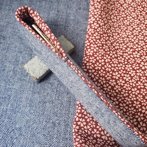 Pode incluir: Um estojo para canetas de tecido azul em espinha de peixe com forro estampado floral vermelho. O estojo está aberto, revelando uma caneta prateada. O fundo apresenta um tecido azul em espinha de peixe e um tecido floral vermelho.