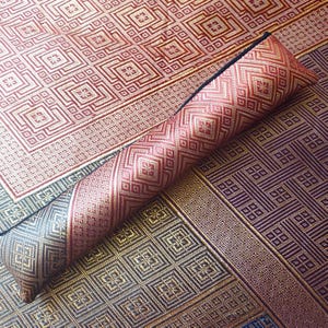 May include: A cylindrical bolster pillow featuring a woven geometric pattern in shades of deep red, gold, and black. The pillow rests diagonally across a flat, rectangular mat that shares the same intricate, repeating square and diamond design.