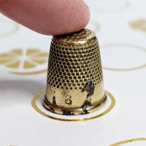 May include: A gold thimble with a textured surface and small holes. The thimble shows some signs of wear and the number "6" is visible. It sits on a white surface decorated with gold citrus fruit and circle designs.