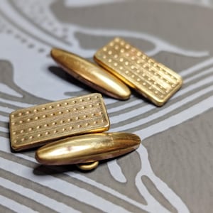 Op de afbeelding: Close-up van twee paar goudkleurige manchetknopen. Elke manchetknop heeft een rechthoekige voorkant met een gestippeld patroon. De manchetknopen liggen op een oppervlak met een patroon, wat duidt op een formeel accessoire.