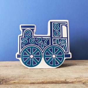 Może przedstawiać: Ozdobny przedmiot w kształcie pociągu z białą podstawą i niebieskimi akcentami. Pociąg ma dwa turkusowe koła i różne niebieskie cyfry. Tło jest niebieskie, a przedmiot spoczywa na drewnianej powierzchni.
