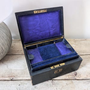 May include: An open, antique black wooden box with a hinged lid. The interior is lined with a vibrant purple fabric, and a smaller tray sits inside. The box has gold-colored metal accents and a keyhole.