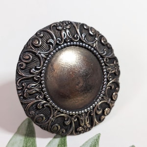 Może przedstawiać: Ozdobny, antyczny guzik z centralnym, metalicznym dyskiem. Guzik ma szczegółowy, wirujący wzór w ciemnym, postarzanym metalicznym wykończeniu, z obramowaniem z koralików. Element dekoracyjny do szycia lub rękodzieła.