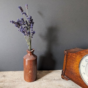 Op de afbeelding: Een bruine keramische vaas met gedroogde lavendel. De vaas heeft een smalle hals en een afgeronde body. Een houten klok met een witte wijzerplaat en zwarte cijfers staat op de achtergrond. De scène speelt zich af op een houten oppervlak tegen een donkergrijze muur.