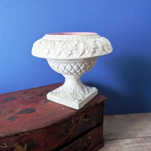 Op de afbeelding: Een witte, sierlijke urn met een gedetailleerd bladerenpatroon rond de rand en de basis. De urn heeft een roze binnenkant en staat op een vierkante basis. De achtergrond is effen blauw en de urn staat op een rode, antieke kist.