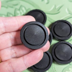 Può includere: Una collezione di tappi di plastica neri e circolari. I tappi hanno un design concentrico in rilievo e sono tenuti in una mano. Lo sfondo è una superficie verde chiaro con un motivo floreale.
