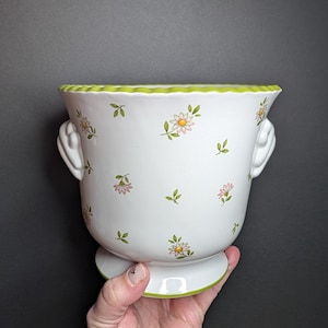 May include: A white ceramic planter featuring a repeating pattern of small pink and yellow flowers with green leaves. The planter has a scalloped lime green rim, a matching lime green base, and two white side handles with a shell-like design.