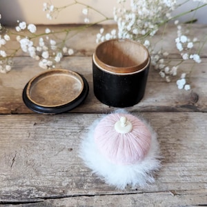 May include: A vintage black and wood powder puff container with a pink puff and white trim. The container is open and the puff is sitting on a wooden surface.