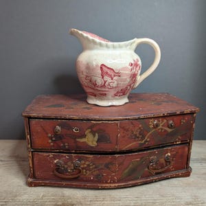 Puede incluir: Una jarra de cerámica roja y blanca vintage con asa y escena pastoril. Se encuentra sobre una pequeña cómoda antigua de madera roja con dos cajones y herrajes ornamentados. El cofre tiene diseños pintados de flores y pájaros.