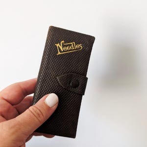 May include: A dark brown rectangular case with a textured surface and a snap button closure. The word Needles is printed in gold lettering on the front flap. The case is constructed from a firm material and features a flap that folds over the body.