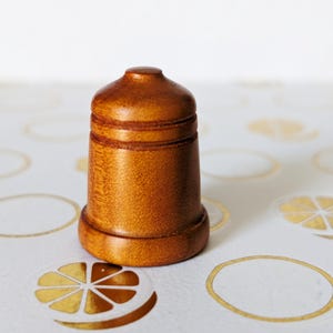 May include: A small, wooden thimble with a rounded top and a flared base. The thimble is a warm, medium brown color and has two carved bands around the middle. It is sitting on a white surface with gold citrus fruit designs.