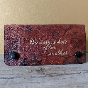 Op de afbeelding: Een rechthoekige bruin lederen portemonnee met de gouden tekst "One darned hole after another". De portemonnee heeft een textuur en twee zwarte drukknopen.