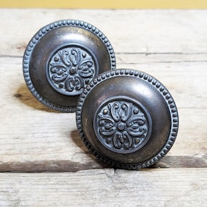 Op de afbeelding: Twee antieke deurknoppen in bronskleur met een bloemmotief in het midden. De knoppen hebben een kralenrand en zijn geplaatst tegen een rustieke houten achtergrond. De knoppen zijn rond en lijken van metaal te zijn.