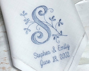 May include: White handkerchief with a blue embroidered initial "S" and floral accents. The text "Stephen & Emily June 18, 2022" is embroidered below the initial. The handkerchief has a stitched border.