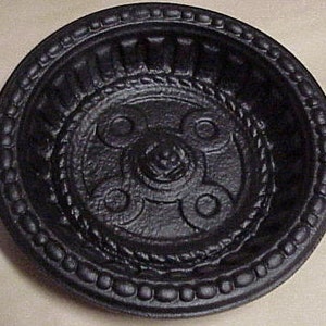 May include: A round, black cast iron dish with an ornate design. The dish features a raised, textured pattern with a central floral motif, rope-like detailing, and a beaded edge. The dish is likely for decorative or functional use.