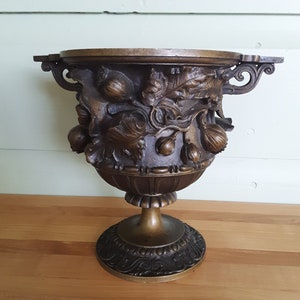 May include: A bronze decorative urn with an ornate design featuring leaves and acorns. The urn has a round base and a wide, open top.