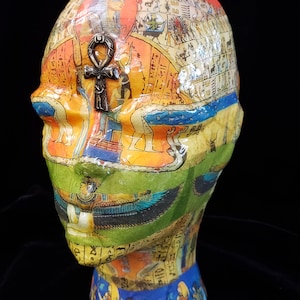 Può includere: Una testa di manichino ricoperta da un collage di immagini colorate a tema egizio, tra cui geroglifici, figure e divinità. Un simbolo ankh metallico è fissato sulla fronte. La superficie ha una finitura lucida con vivaci sezioni arancioni, verdi e blu.